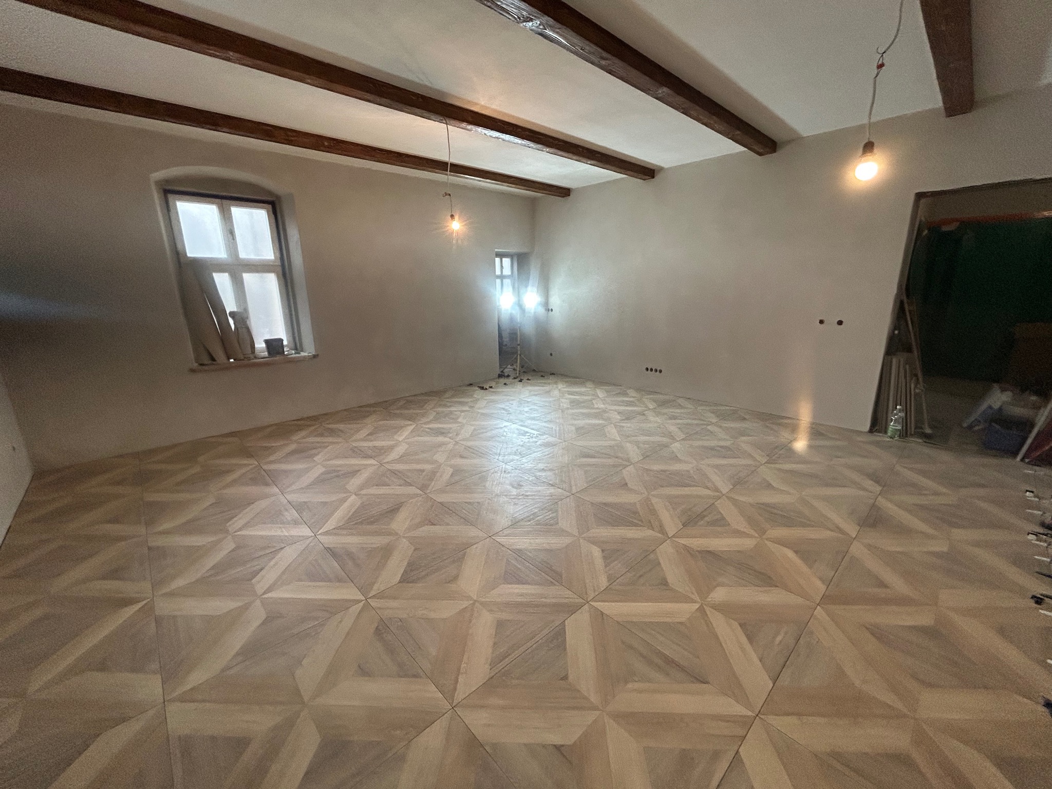 Ukończona podłoga z parkietu w geometryczny wzór w przestronnym, surowym wnętrzu z drewnianymi belkami stropowymi i oknem. Widoczne ślady prac wykończeniowych.