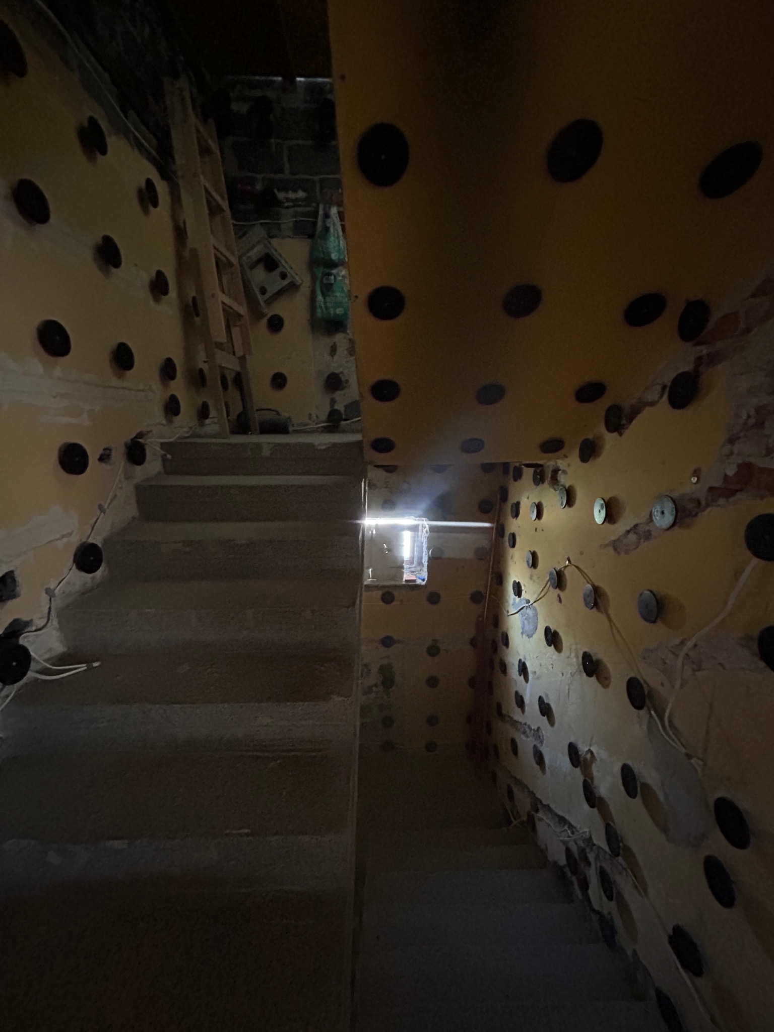 Schody betonowe w trakcie prac wykończeniowych, ściany z żółtą izolacją i czarnymi mocowaniami. Widoczne przewody i fragment drabiny.