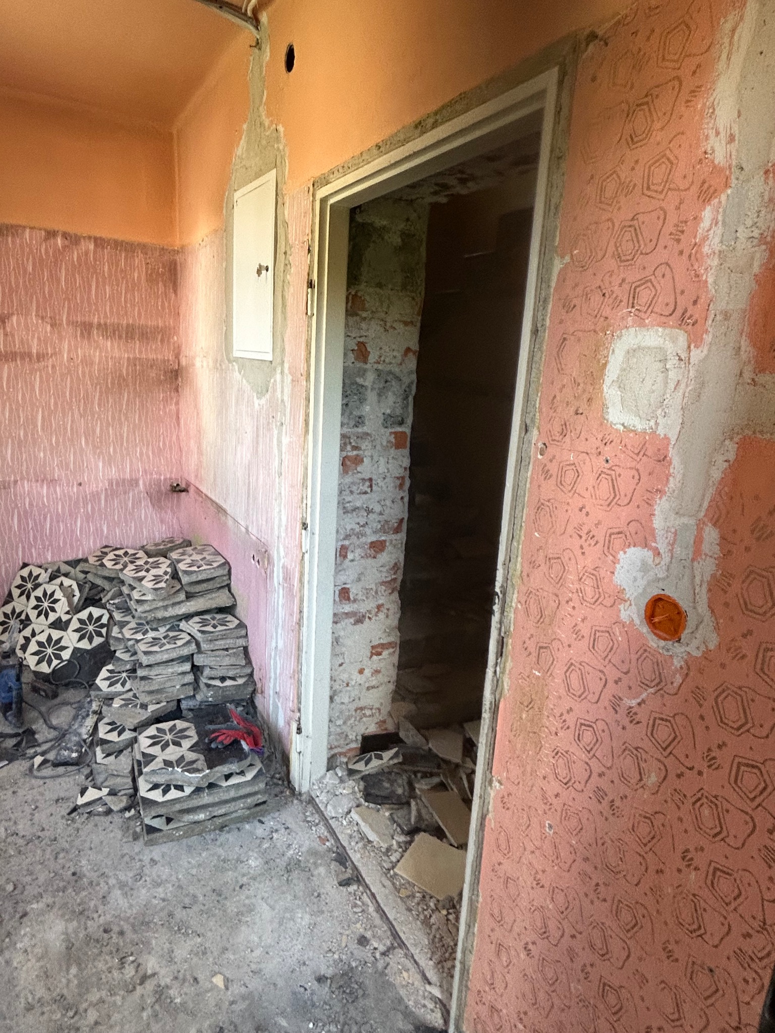 Demontaż starej glazury w łazience. Stos płytek z motywem kwiatowym, ściany z resztkami tapety i kleju, widoczne ślady po instalacjach. Przygotowanie do remontu.