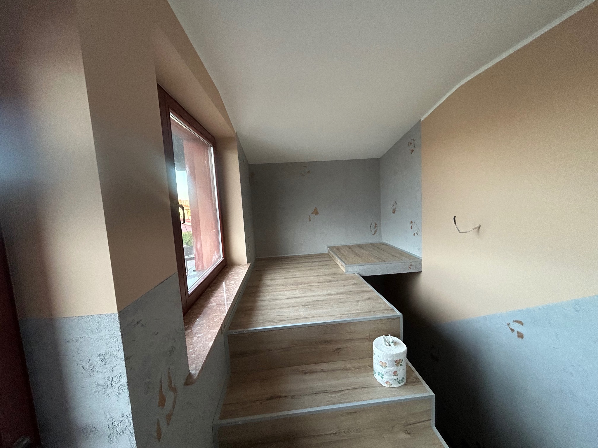 Wnętrze po remoncie: schody z drewnopodobnych paneli prowadzą na podest, ściany częściowo pomalowane na beżowo, częściowo z szarą strukturą, okno z brązową ramą.