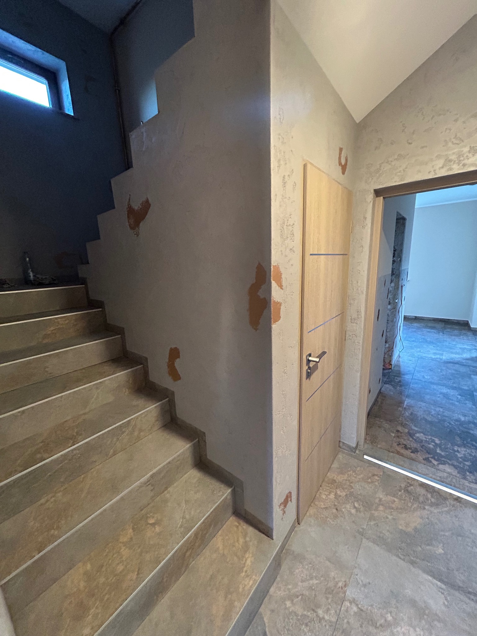 Wnętrze w trakcie wykańczania: schody z płytek, ściany z fakturą, drewniane drzwi. Widoczne niedociągnięcia w postaci plam na ścianie.