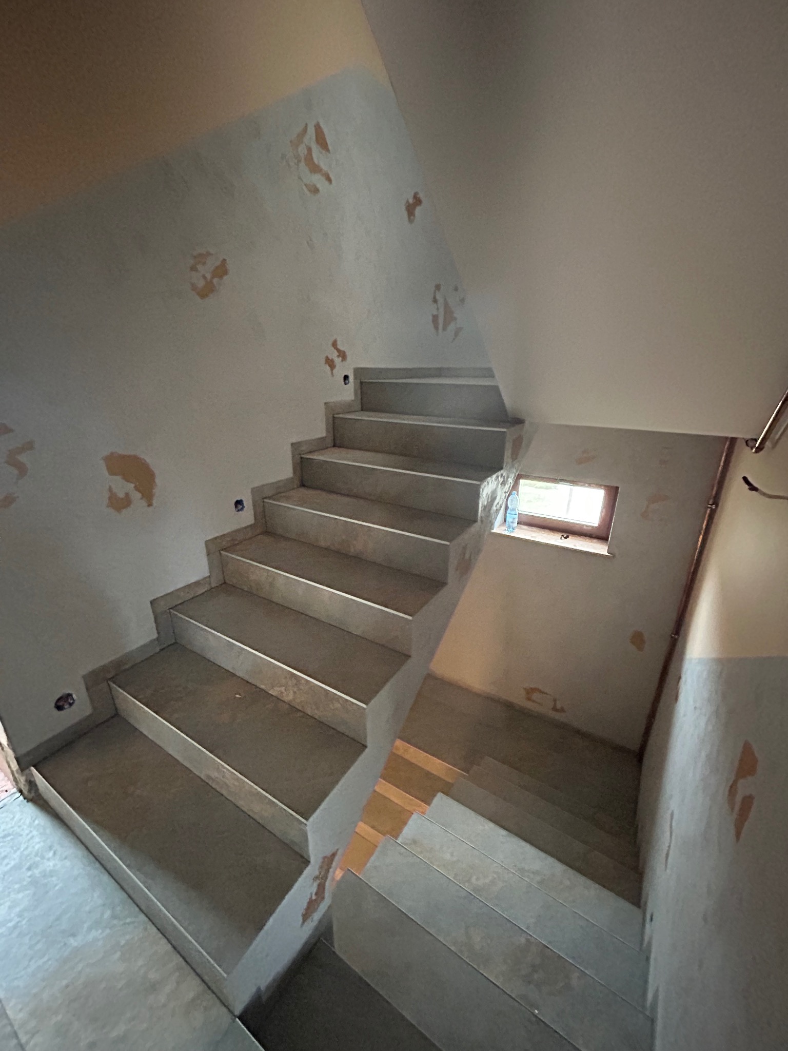 Schody w trakcie remontu, widoczne ślady po starym wykończeniu ścian, surowe stopnie schodów, małe okno na podeście.