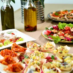 Dekoracyjnie podane przystawki w drewnianych łódeczkach na białym talerzu, obok butelki z oliwą i papryczkami chili oraz dania z łososiem i warzywami.