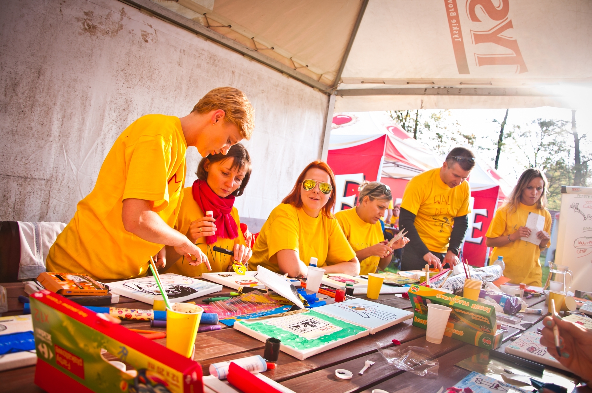 Grupa osób w żółtych koszulkach pracuje twórczo przy stołach na zewnątrz, malując obrazy na płótnach podczas imprezy integracyjnej. Na stołach widoczne farby, pędzle, kubki i inne materiały...