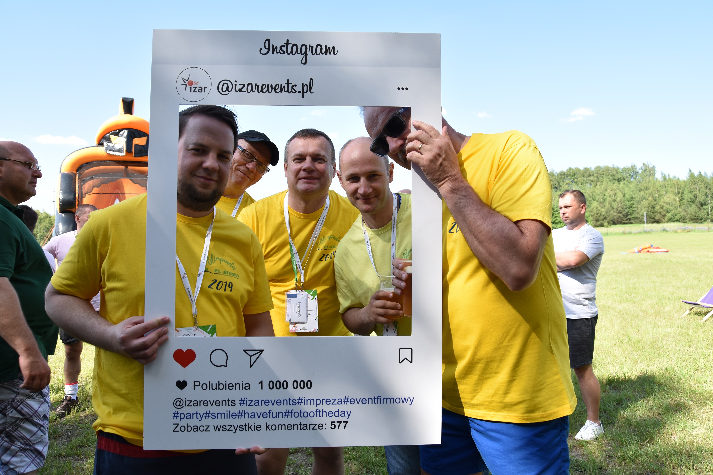 Grupa mężczyzn w żółtych koszulkach pozuje do zdjęcia w ramce imitującej Instagram na imprezie plenerowej w Katowicach, w tle dmuchany zamek i zielona trawa.