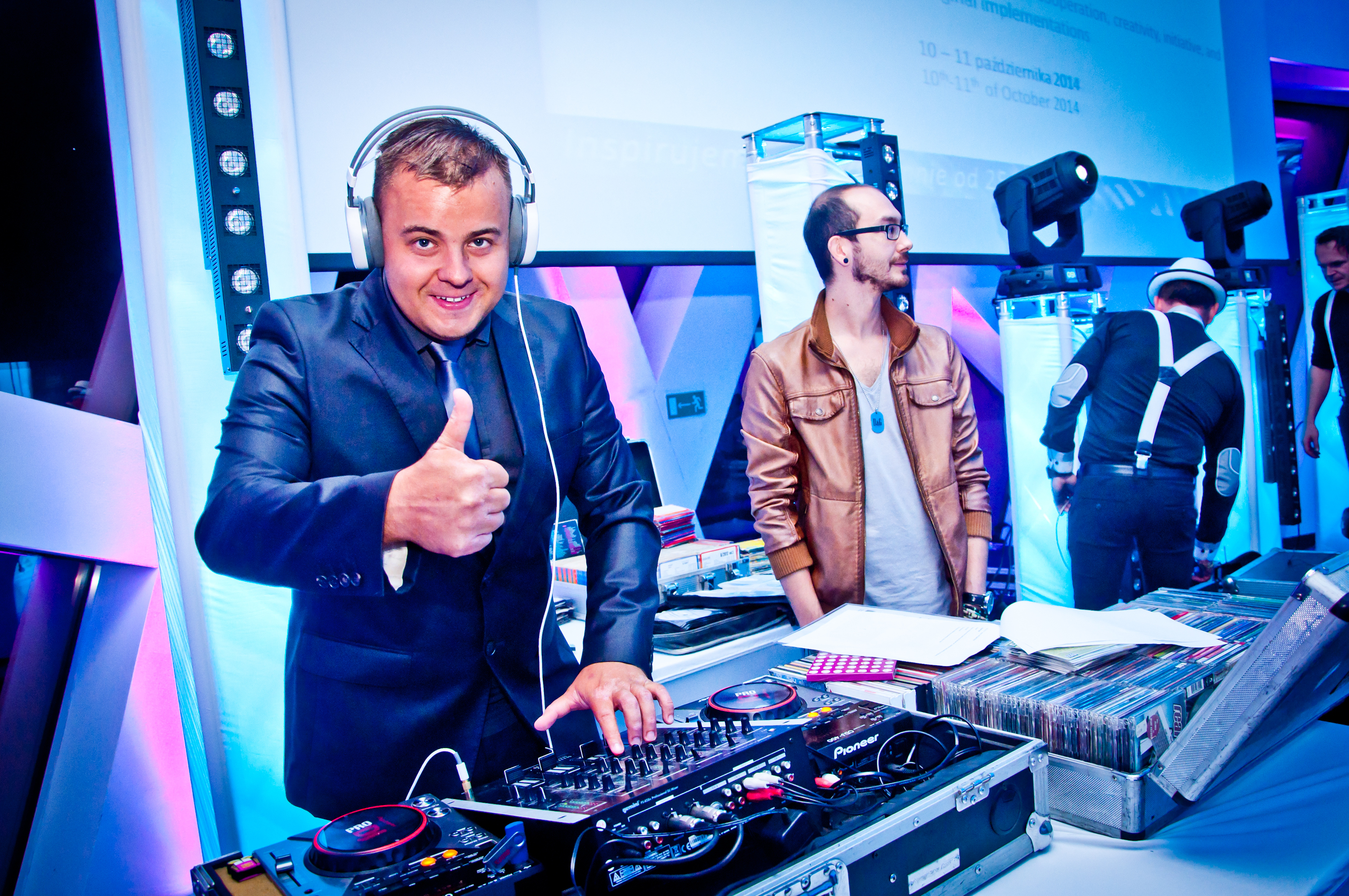 Uśmiechnięty DJ w garniturze i słuchawkach, z uniesionym kciukiem, miksuje muzykę na profesjonalnym sprzęcie Pioneer na tle oświetlonej sceny z innymi osobami.