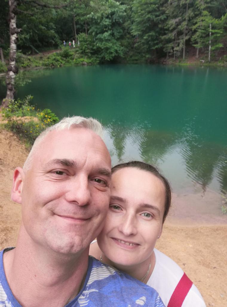 Selfie pary na tle turkusowego jeziora otoczonego zielenią. Uśmiechnięci, blisko siebie, z widocznym odbiciem drzew w wodzie. Jasne, naturalne światło.