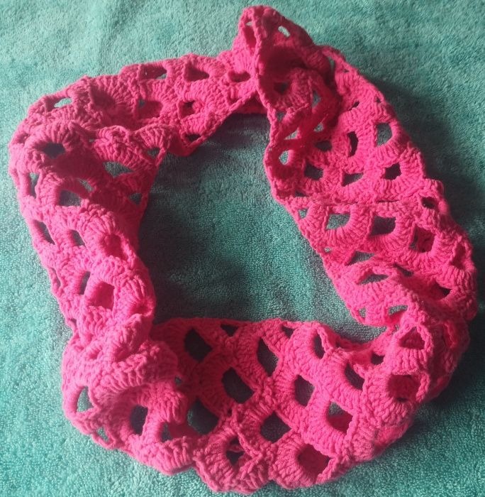 Różowy, ażurowy szal dziergany na drutach, ułożony w okrąg na turkusowym tle.