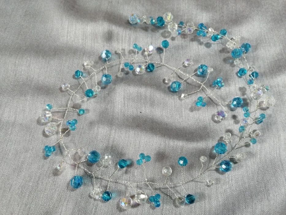 Delikatna ozdoba do włosów z srebrnego drutu, turkusowych i przezroczystych kryształków, leżąca na srebrzystoszarym tle.