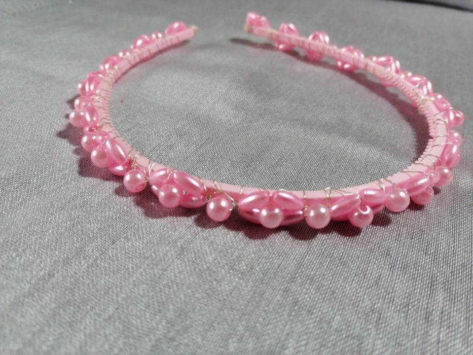 Różowa opaska do włosów, ozdobiona perłami i owinięta srebrnym drucikiem, leżąca na szarym tle.