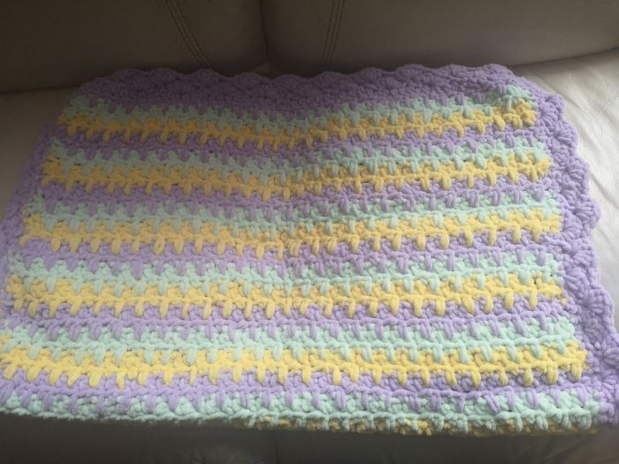 Ręcznie robiony kocyk dla dziecka w pastelowych odcieniach fioletu, żółci i zieleni, z dekoracyjnym brzegiem, leżący na skórzanej sofie.