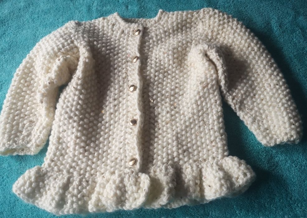 Kremowy, ręcznie robiony sweterek dziecięcy z falbanką na dole i złotymi akcentami, zapinany na cztery guziki, leżący na turkusowym tle.