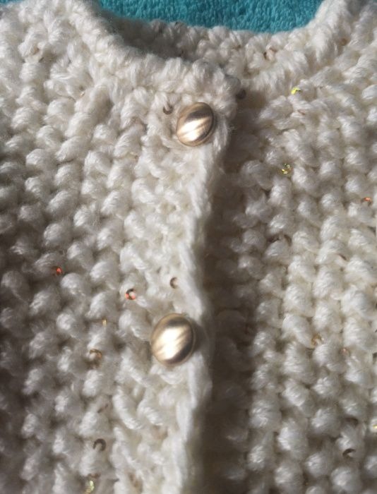 Kremowy, ręcznie robiony kardigan z grubego splotu, ozdobiony złotymi cekinami i dwoma błyszczącymi, złotymi guzikami.