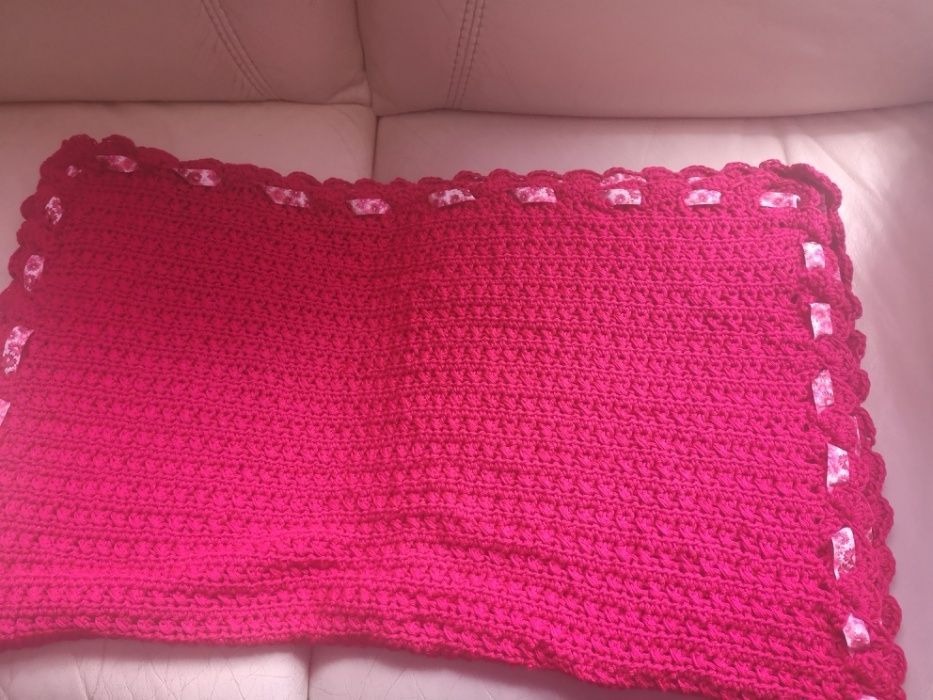 Ręcznie robiony, intensywnie różowy kocyk z włóczki, wykończony ozdobną falbanką i wstążeczkami w kwiatowy wzór, leżący na jasnej skórzanej kanapie.