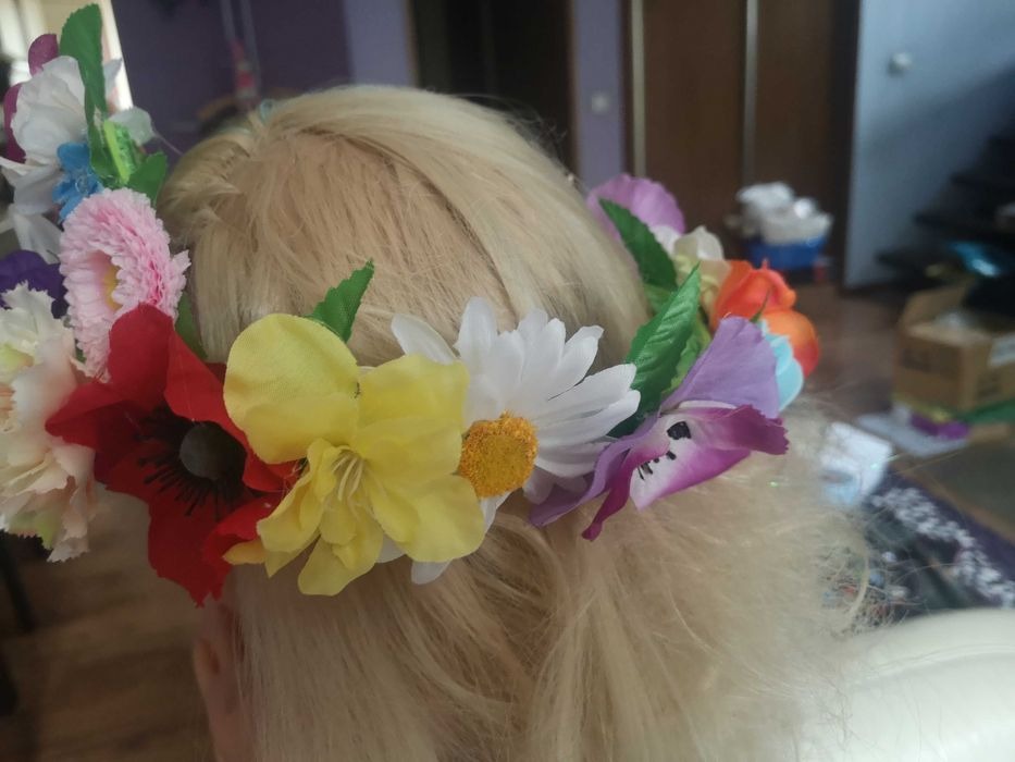 Blond włosy z wiankiem z różnokolorowych, sztucznych kwiatów, w tle widoczne elementy wyposażenia wnętrza.