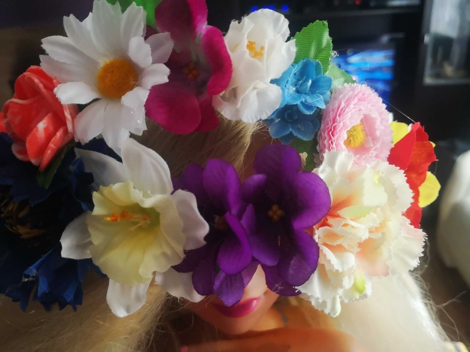 Lalka z blond włosami i wiankiem z różnokolorowych sztucznych kwiatów: białe margaretki, fioletowe fiołki, czerwone tulipany, niebieskie niezapominajki, różowe goździki i żółte żonkile.