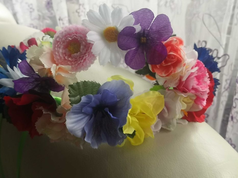 Wianek z różnokolorowych, sztucznych kwiatów, w tym bratków, astrów i margerytek, leżący na jasnej, gładkiej powierzchni, z zasłoną w tle.