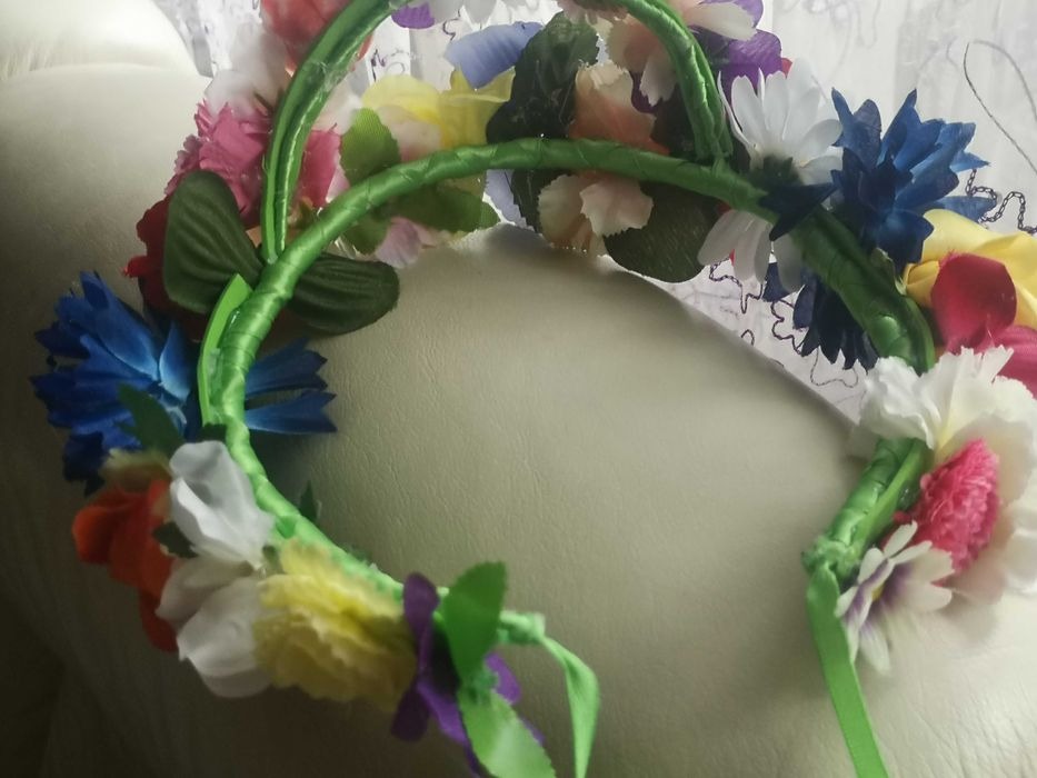 Ręcznie robiony wianek z kolorowych, sztucznych kwiatów na zielonej, owiniętej tasiemką obręczy, leżący na jasnej powierzchni z koronkowym tłem.
