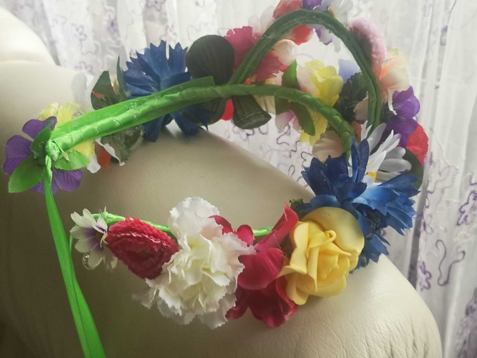 Ręcznie robiony wianek z kolorowych, sztucznych kwiatów na zielonej, satynowej opasce, leżący na jasnym tle.