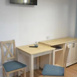 Pokój hotelowy z drewnianym stołem, krzesłami z niebieskimi siedziskami, telewizorem na ścianie i komodą w jasnym drewnie.