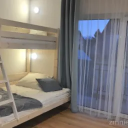 Jasne drewniane łóżko piętrowe z drabinką w pokoju z oknem zasłoniętym firanką, widok na dach budynku.