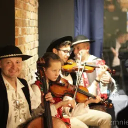 Zespół muzyczny w strojach ludowych grający na skrzypcach i wiolonczeli, występ na tle ceglanej ściany i niebieskiej zasłony.