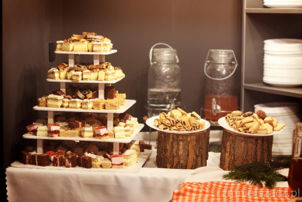 Bogato zastawiony stół z paterą pełną różnorodnych ciast i ciasteczek, napoje w szklanych słojach z kranikami, dekoracja z plastrów drewna i gałązki świerku.