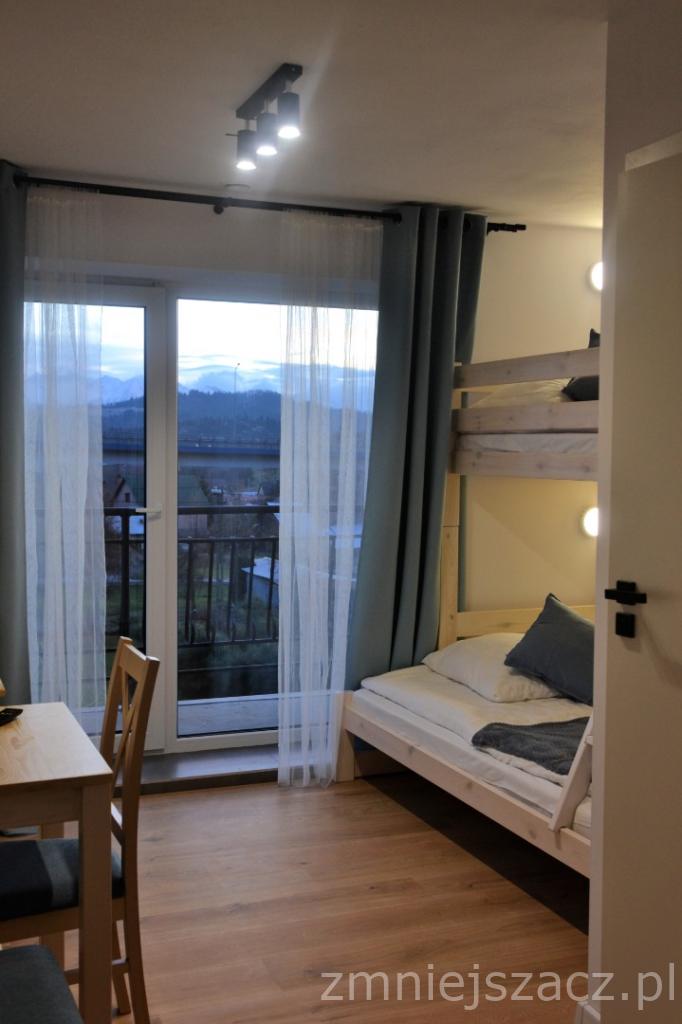 Pokój z łóżkiem piętrowym, biurkiem i widokiem na góry za oknem balkonowym z firankami i zasłonami.