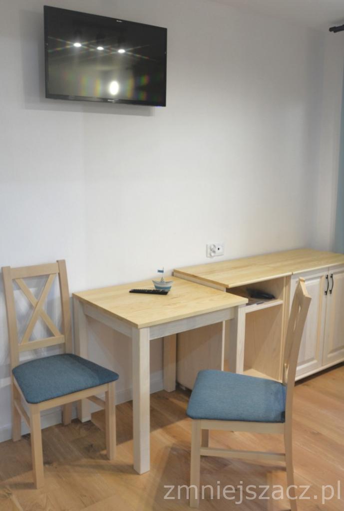 Pokój hotelowy z drewnianym stołem, krzesłami z niebieskimi siedziskami, telewizorem na ścianie i komodą w jasnym drewnie.