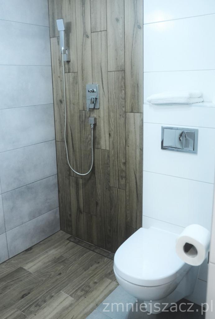 Nowoczesna łazienka z kabiną prysznicową wyłożoną płytkami imitującymi drewno, białą toaletą i ręcznikiem na półce.