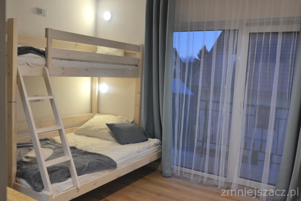 Jasne drewniane łóżko piętrowe z drabinką w pokoju z oknem zasłoniętym firanką, widok na dach budynku.