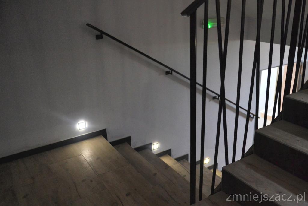 Klatka schodowa z drewnianymi schodami, oświetleniem punktowym w ścianie i czarną, nowoczesną balustradą z pionowych prętów.