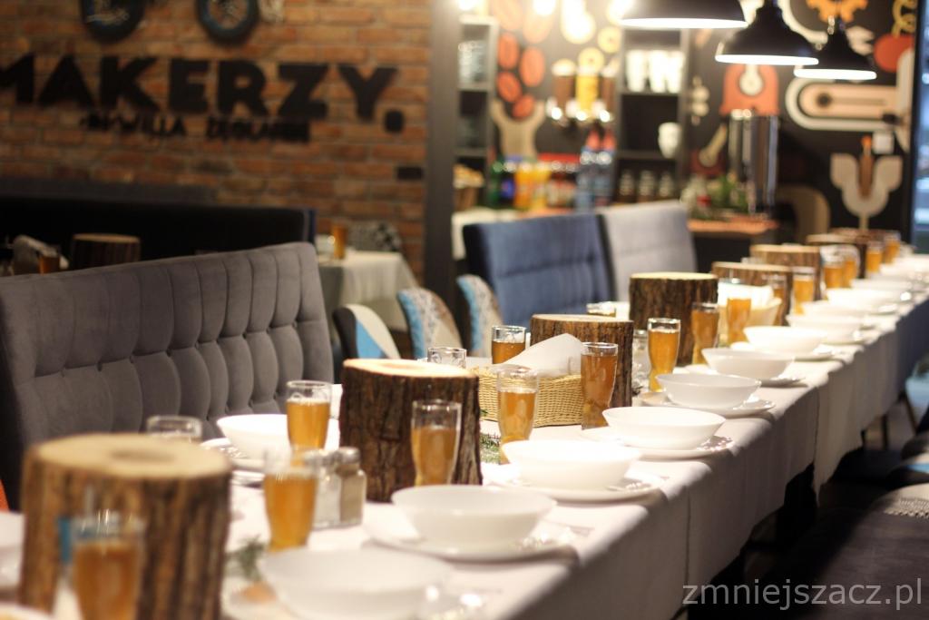 Długi stół nakryty białym obrusem, z białymi miseczkami, szklankami z napojem i dekoracjami z pniaków drewna, przygotowany na imprezę w restauracji z ceglaną ścianą i napisem 'MAKERZY'.