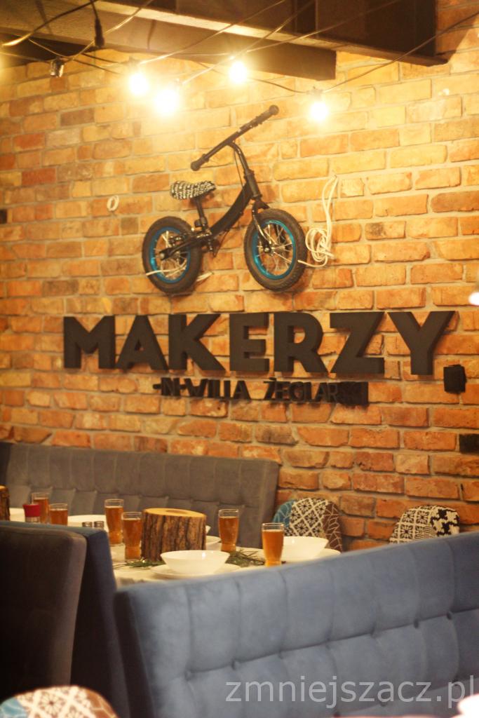 Wystrój wnętrza restauracji z ceglaną ścianą, rowerem na ścianie i napisem 'MAKERZY - VILLA ŻEGLARSKA' nad stołem z nakryciami i napojami.