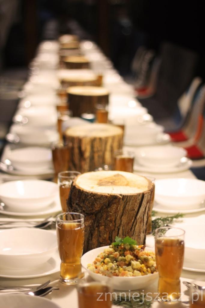 Długi stół z białymi naczyniami, szklankami z brązowym napojem i dekoracjami z pniaków drewna oraz miseczkami z sałatką warzywną, przygotowany na przyjęcie.