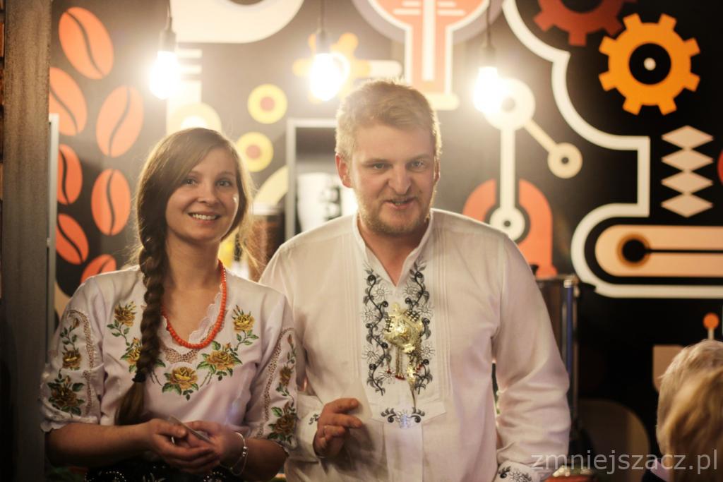 Uśmiechnięta para w tradycyjnych strojach ludowych na tle dekoracji eventowej z motywami kawy i mechaniki.