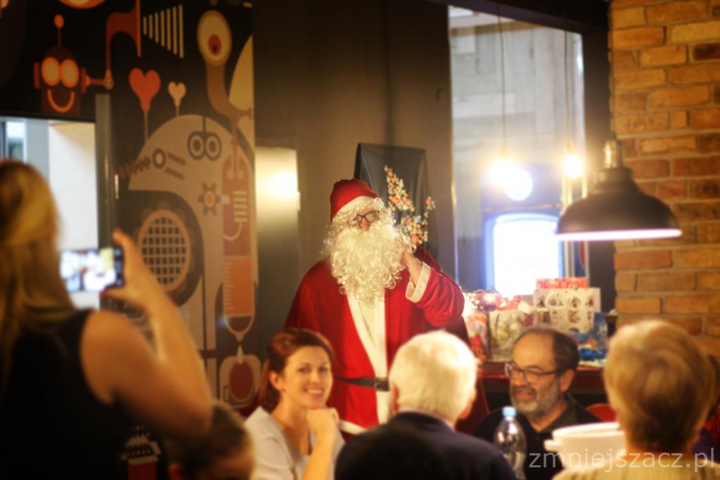 Mikołaj w czerwonym stroju i okularach stoi przed grupą osób siedzących przy stole, w tle dekoracje i prezenty, osoba robiąca zdjęcie telefonem.