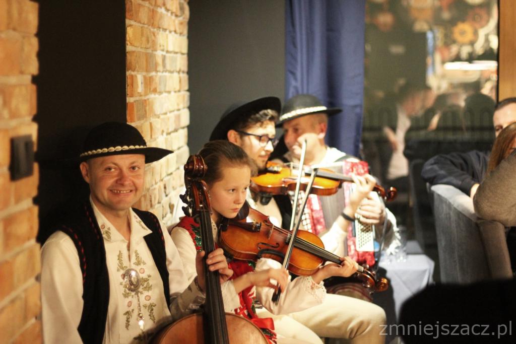 Zespół muzyczny w strojach ludowych grający na skrzypcach i wiolonczeli, występ na tle ceglanej ściany i niebieskiej zasłony.