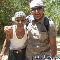 Uśmiechnięty turysta w okularach słonecznych obejmuje starszego mężczyznę w tradycyjnym nakryciu głowy, trzymającego koszyk z datkami na tle zieleni.