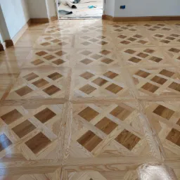 Ułożony parkiet w geometryczny wzór z jasnego i ciemnego drewna, widoczne listwy przypodłogowe i otwarte drzwi do kolejnego pomieszczenia z materiałami budowlanymi.