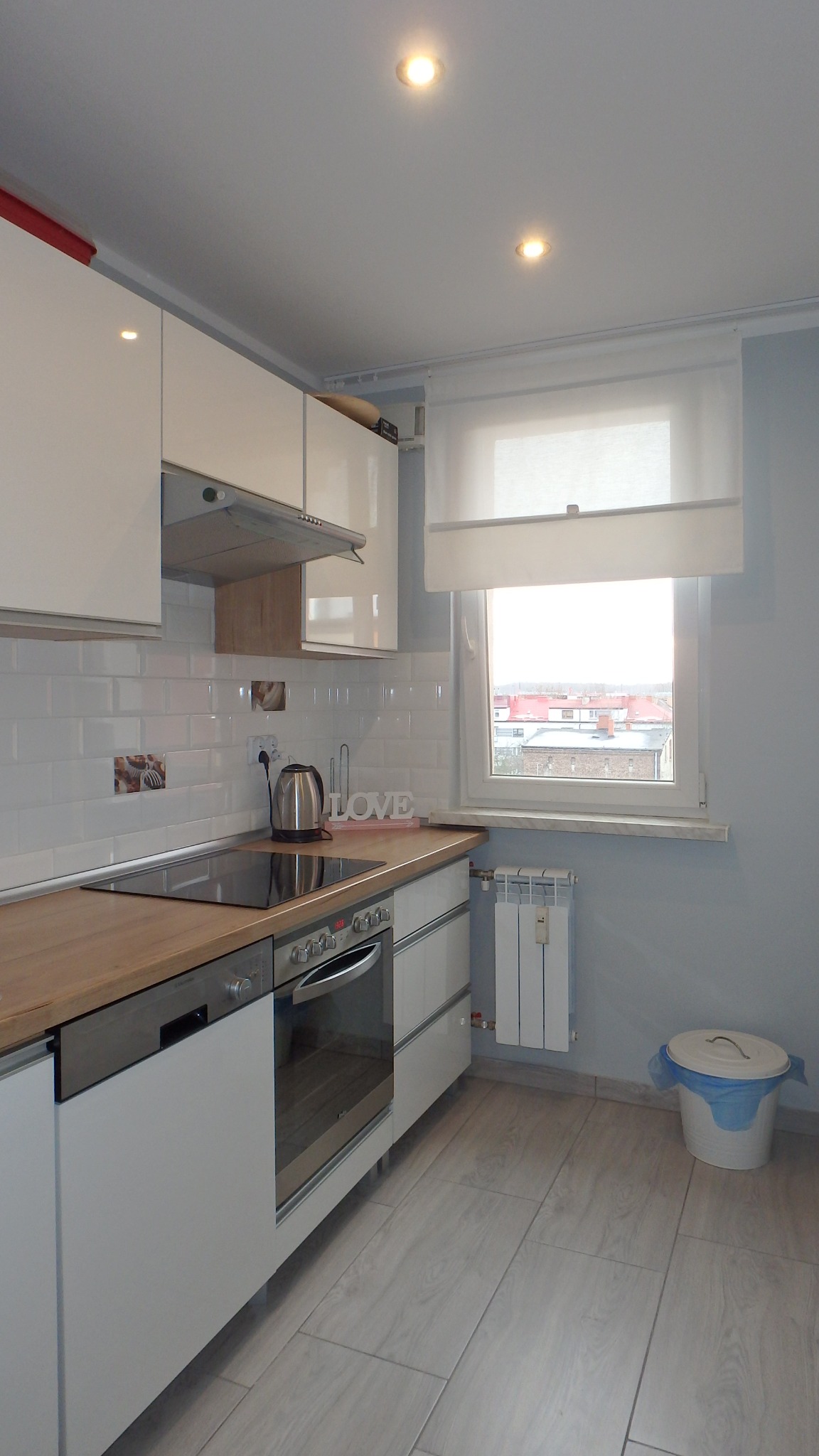 Jasna kuchnia z białymi szafkami, drewnianym blatem, płytą indukcyjną i piekarnikiem. Na blacie czajnik elektryczny i dekoracja z napisem LOVE. Widoczne okno z roletą rzymską.