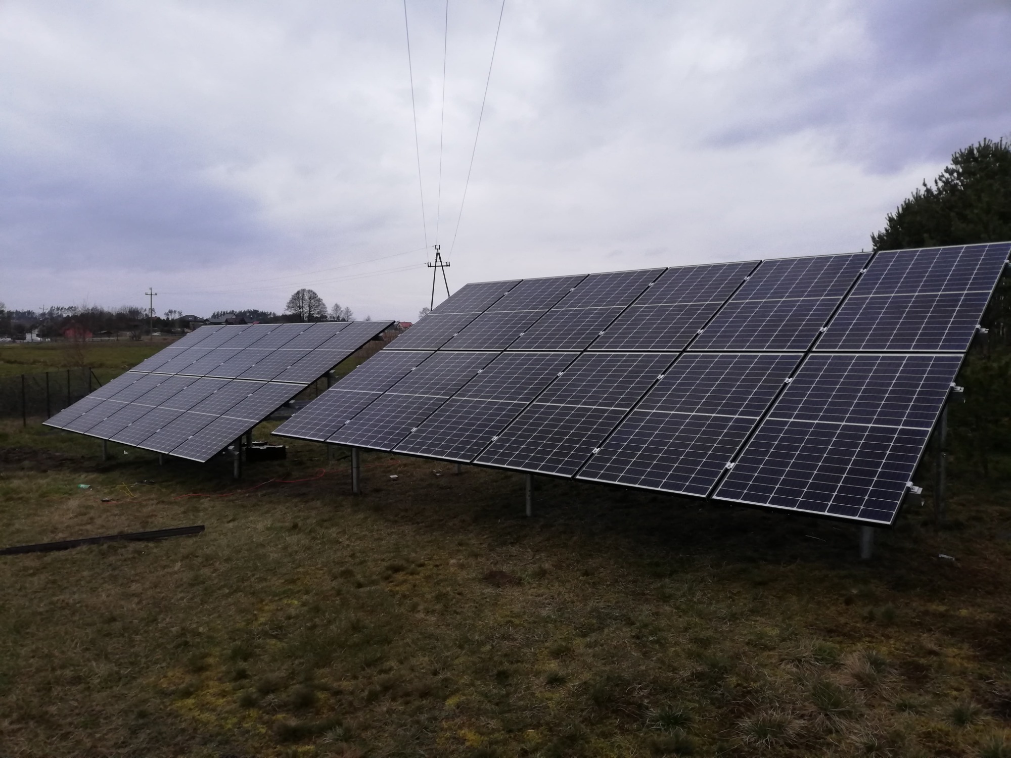 Dwa rzędy paneli słonecznych na trawiastym terenie w Gdańsku, widoczne słupy energetyczne i pochmurne niebo.