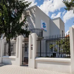 Elewacja budynku kliniki z logo, ogrodzona czarnym metalowym płotem na tle błękitnego nieba z chmurami.