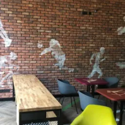 Wnętrze kawiarni z ceglaną ścianą i sylwetkami sportowców, drewniany blat stołu z metalową siatką, żółte i szare krzesła oraz okrągły stolik kawowy.