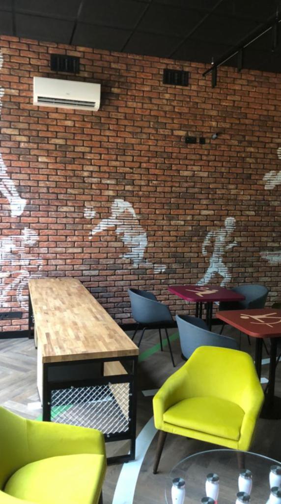 Wnętrze kawiarni z ceglaną ścianą i sylwetkami sportowców, drewniany blat stołu z metalową siatką, żółte i szare krzesła oraz okrągły stolik kawowy.