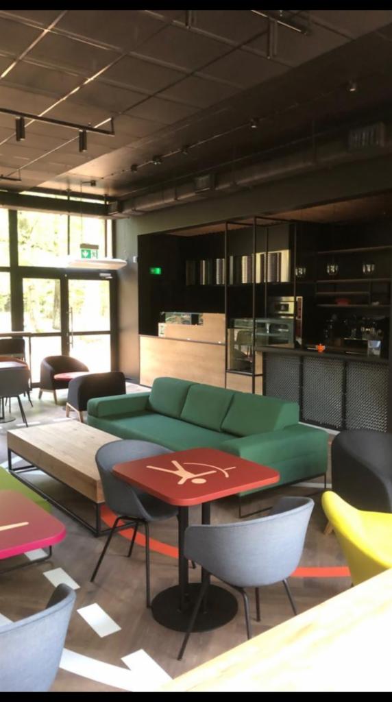 Wnętrze kawiarni z zieloną sofą, stolikami z czerwonymi blatami z białym symbolem figury ludzkiej, szarymi krzesłami i drewnianym stolikiem kawowym na tle czarnego baru z metalową siatką...