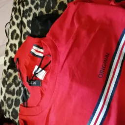 Czerwona koszulka z napisem 'ORIGINAL' i paskami w kolorach białym i granatowym, leżąca na stercie ubrań z widoczną metką H&M.