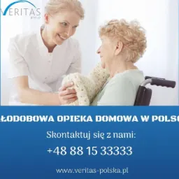 Uśmiechnięta opiekunka w białym uniformie rozmawia z starszą kobietą na wózku inwalidzkim, okrytą wełnianym pledem.