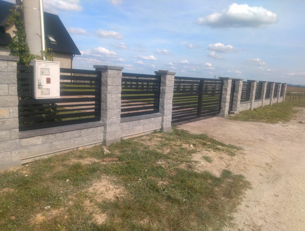 Nowoczesne ogrodzenie panelowe z szarego kamienia i czarnych poziomych przęseł, widok z zewnątrz posesji, na pierwszym planie trawa i gruntowa droga.