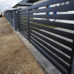 Nowoczesna, pozioma brama przesuwna z czarnych, metalowych profili, z widocznym fragmentem posesji i osobą w tle, zamontowana na szarej kostce brukowej.